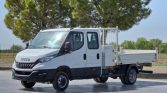 White Iveco flatbed utility truck with dual cab, parked on a paved surface with trees in the background