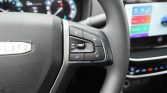 Close-up of a car steering wheel with audio/phone control buttons and a dashboard screen in the background.