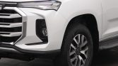 Close-up of a white pickup truck's front left corner, highlighting the headlight, grille, and large alloy wheel with tire visible.