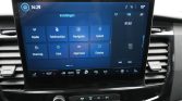 Infotainment touchscreen showing settings tiles (Radio, Telefoonlijst, Navigatie, Geluid, Voertuig, Klok, Algemeen, Display) with climate controls below in a car cabin.