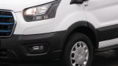 Front-left corner of a white van showing the headlight, grille with blue accents, and front wheel on a dark surface.