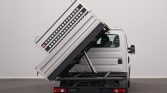 White Iveco Daily dump truck with its metal bed raised at a steep angle via hydraulic lift, ready to unload.