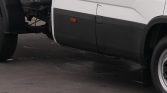 Side view of a white commercial truck with a black lower body panel and exposed wheel; a circular manhole cover is on the pavement nearby.