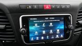 Car dashboard center console with a touchscreen displaying audio, media, settings, compass, radio, and backup camera icons.