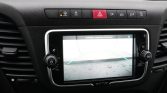 Dashboard infotainment screen showing a car rearview camera display with blue guiding lines on a wet surface; controls surround the screen.