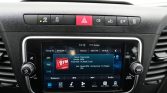 Car infotainment touchscreen showing a blue radio interface with presets and icons, flanked by round knobs on a black dashboard.