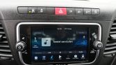 Infotainment touchscreen in a car console showing media controls and time.
