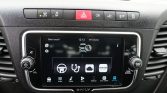 Center console of a car showing a touchscreen infotainment system with app icons and menus, flanked by volume and tune knobs on black dashboard