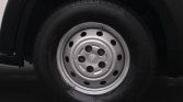 Close-up of a car wheel with a silver steel rim, five lug nuts, and circular holes, center cap featuring a logo ( Peugeot-style ).