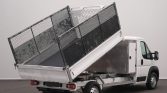 White pickup-style dump truck with its bed raised, unloading a tilted metal cargo cage into the back.