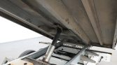 Underside view of a trailer with its hydraulic landing gear leg extended, showing the metal frame and wheel in the background