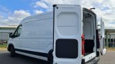 White cargo van with its rear doors open, revealing a spacious empty loading area and black rear interior panels on a paved lot under a partly cloudy sky