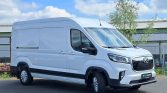 White cargo van parked on a paved lot beside an industrial building, front-right view showing large grille and black bumper edges in daylight.