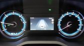 Electric vehicle instrument cluster: blue-lit left speed dial and right speedometer with central touchscreen showing lane-keeping guidance and a car icon inside the road lanes.