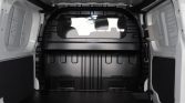 Empty white cargo van interior with black paneling and a windowed bulkhead seen from the rear doors open, front seats visible through the window.