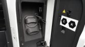 Close-up of an electric vehicle charging inlet inside an open panel, with two circular caps and a coiled charging cable visible.