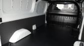 Interior of an empty white cargo van with a black non-slip floor, wheel arches, and a partition with a rear window at the front.