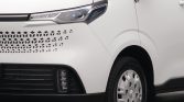 Front-left view of a white van showing a decorative hexagonal grille, slim headlight, and front wheel.