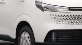 Front-left view of a white van showing the headlight, perforated grille pattern, and front wheel edge.