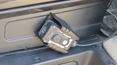 Portable car battery tester/diagnostic device with two round buttons, resting on a vehicle interior panel and connected by a cord.