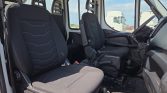 Interior of a truck cab showing two black fabric seats, steering wheel, gear lever, and pedals with a view through large windows to parked trucks outside