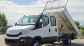 White Iveco dump truck with its bed raised for dumping, parked on a paved lot under a blue sky.