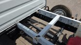 Rear view of a utility trailer frame showing the axle, leaf springs, and wheel, with a white metal frame and rust spots on the joints.