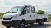 White Iveco flatbed truck with double cab, parked on a paved lot under a blue sky, cargo bed at rear.
