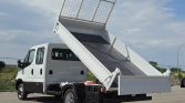 White utility/dump truck with a tilted hydraulic bed raised, in an open lot under a cloudy sky