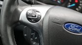 Close-up of a Ford steering wheel with a circular multi-function pad showing OK and directional arrows on the left side of the wheel.