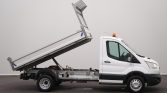 White utility truck with a raised tilt-bed ready to dump cargo, side view with orange beacon on cab roof and exposed chassis beneath the bed