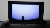 In-dash car display showing the backup camera view of a blue-toned parking area with a small dark object near the bumper in the center.
