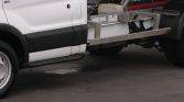 White tow truck with a metal flatbed chassis and hydraulic lift visible behind the cab, on pavement.