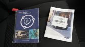 Two Ford vehicle manuals on a car seat: a blue BlueOval Charge Network pamphlet and a white Transit owner's manual with a barcode sticker.