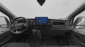 Front cockpit of a vehicle showing a large center touchscreen, steering wheel, and gray dashboard.