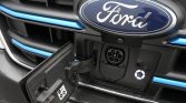 Front of a Ford vehicle with a circular charging port open, showing a power socket and blue accent trims on the grille area.