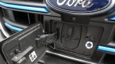 Close-up of a Ford front end with the charging port door open, exposing the inlet beneath the bumper and blue accent trim.