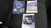 Three Ford vehicle manuals on a car seat: a blue Charge Network booklet on the left, a white road-scene brochure on the top right, and a Transit owner’s guide with a van on the bottom.