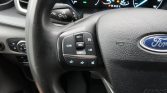 Steering wheel close-up showing Ford emblem and left-side cruise control buttons (CAN, SET+, RES, SET-, LIM) with volume/mute icons; illuminated dashboard gauges in the background.