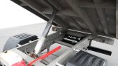 Industrial hydraulic lift mechanism under a truck bed, showing a gas can, red bar, and metal support frame.