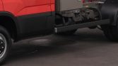 Red commercial truck with a long black side step extending beneath the cab and exposed mechanical components in the chassis area.