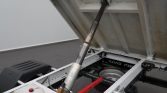 Dump truck bed raised on a hydraulic piston; view of white chassis, red bar, and utility components underneath.