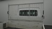 White van visible behind a metal wire-mage cargo barrier in a flatbed truck bed, showing through the grid window.
