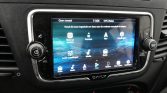 In-car infotainment touchscreen showing the home screen with icons (Back-up camera, Kompas, Nav, Instellingen, Audio instellingen, Media, Radio, Apps, Telefoon) on a blue abstract background; left volume knob and right tuning knob visible.