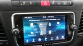 Car infotainment touchscreen in a car dashboard showing navigation, media, and settings icons with a volume knob nearby.