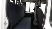 Interior of a white van showing a dark fabric three-seat bench along the left wall, red seatbelt latches, and a front passenger seat; door open on left side.