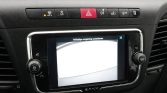 Car dashboard center console with a touchscreen display showing a camera view and the Dutch prompt 'Volledige omgeving controleren' above the image, flanked by volume and tuner controls and a red hazard button above the screen.