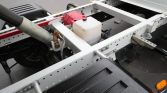 Close-up of a truck trailer chassis showing a hydraulic piston, a white plastic reservoir, and mounting brackets for a tilt/lift mechanism.