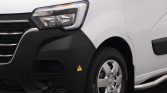 Front-left corner of a white van, showing the headlight, black bumper, and front wheel with a silver rim and tire. A small yellow warning sticker is on the bumper.