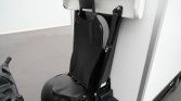 Close-up of a black mesh seat with backrest and safety belt mounted on a small transport vehicle, with a handlebar visible on the left and a white exterior panel behind.